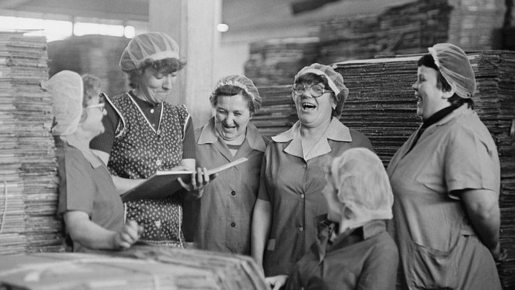 schwar-weiß bild: 5 Frauen stehen inmitten von Stapeln an gedruckten Zeitungen.